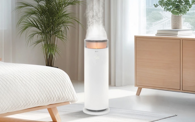 침실에 배치된 LG 퓨리케어 오브제컬렉션 하이드로에센셜 제품이 있으며, 포근 가습 모드가 켜져 있고 수증기가 나오고 있습니다. 창문으로 자연광이 들어오며 녹색 식물이 놓여 있고, 침대는 흰색 침구와 주황색 쿠션으로 꾸며져 있습니다. 작은 탁자와 램프가 보이며, 나무 서랍장 위에 화분이 놓여 있습니다.