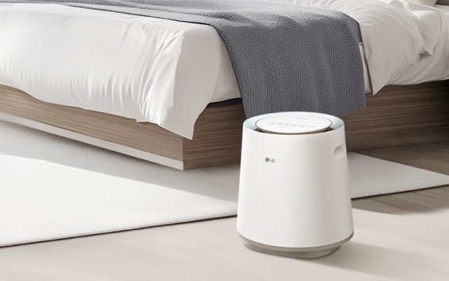 침실에 놓인 LG 퓨리케어 자연 기화 가습기로, 흰색 침구와 베개, 나무 질감의 침대 프레임 옆 흰색 러그 위에 놓여 있습니다.