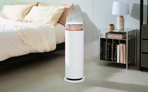 침실 옆 흰색의 LG 퓨리케어 하이드로에센셜 가습기가 작동 중이며, 가습기 상단에서 수증기가 분사되고 있고, 옆에는 탁자 위에 램프, 책, 작은 화분이 놓여있습니다.