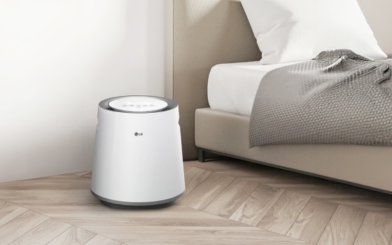 침실에 놓인 흰색 LG 퓨리케어 자연기화 가습기가 보입니다. 가습기는 원통형 디자인에 상단에 제어판이 있습니다. 침대는 베이지색 헤드보드와 흰색 침구가 있으며, 회색 담요가 놓여 있습니다. 방은 흰색 벽과 밝은 나무 바닥으로 꾸며져 있습니다.