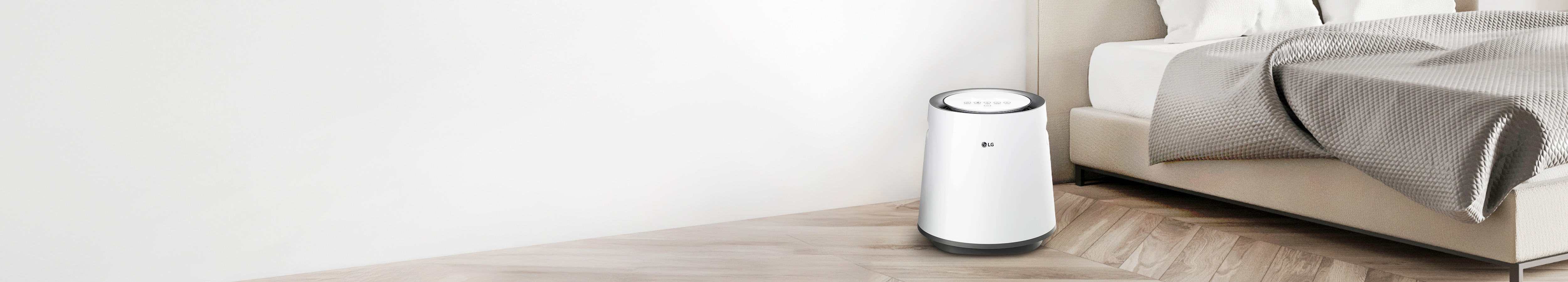 침실에 놓인 흰색 LG 퓨리케어 자연기화 가습기가 보입니다. 가습기는 원통형 디자인에 상단에 제어판이 있습니다. 침대는 베이지색 헤드보드와 흰색 침구가 있으며, 회색 담요가 놓여 있습니다. 방은 흰색 벽과 밝은 나무 바닥으로 꾸며져 있습니다.