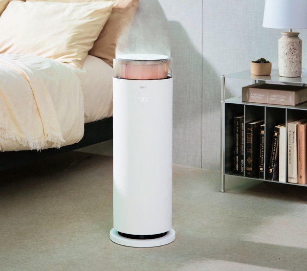 침실 옆 흰색의 LG 퓨리케어 하이드로에센셜 가습기가 작동 중이며, 가습기 상단에서 수증기가 분사되고 있고, 옆에는 탁자 위에 램프, 책, 작은 화분이 놓여있습니다.