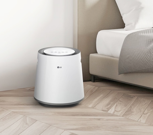 침실에 놓인 흰색 LG 퓨리케어 자연기화 가습기가 보입니다. 가습기는 원통형 디자인에 상단에 제어판이 있습니다. 침대는 베이지색 헤드보드와 흰색 침구가 있으며, 회색 담요가 놓여 있습니다. 방은 흰색 벽과 밝은 나무 바닥으로 꾸며져 있습니다.