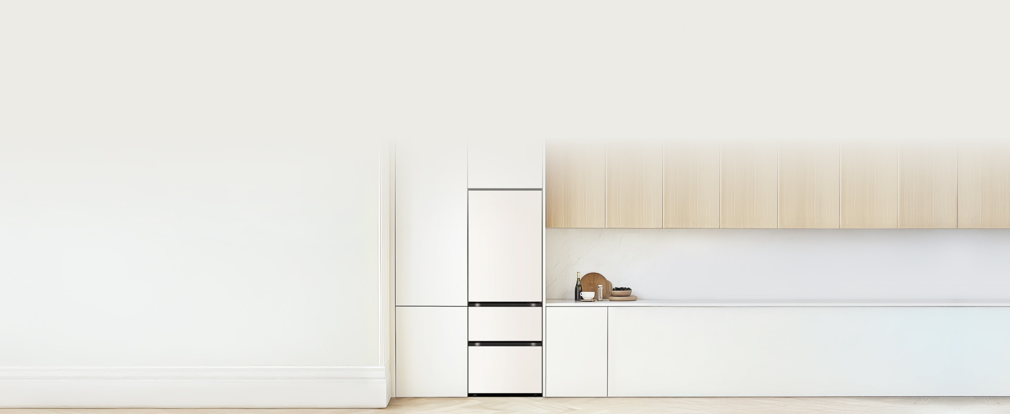 빌트인 구조의 매트 화이트 김치냉장고가 설치되어 있습니다. 그 오른쪽에는 흰색 하부장과 연한 원목 상부장이 조화를 이루고 있으며, 조리대 위에는 와인 병, 컵, 그릇, 나무 도마 등이 깔끔하게 배치되어 있습니다.