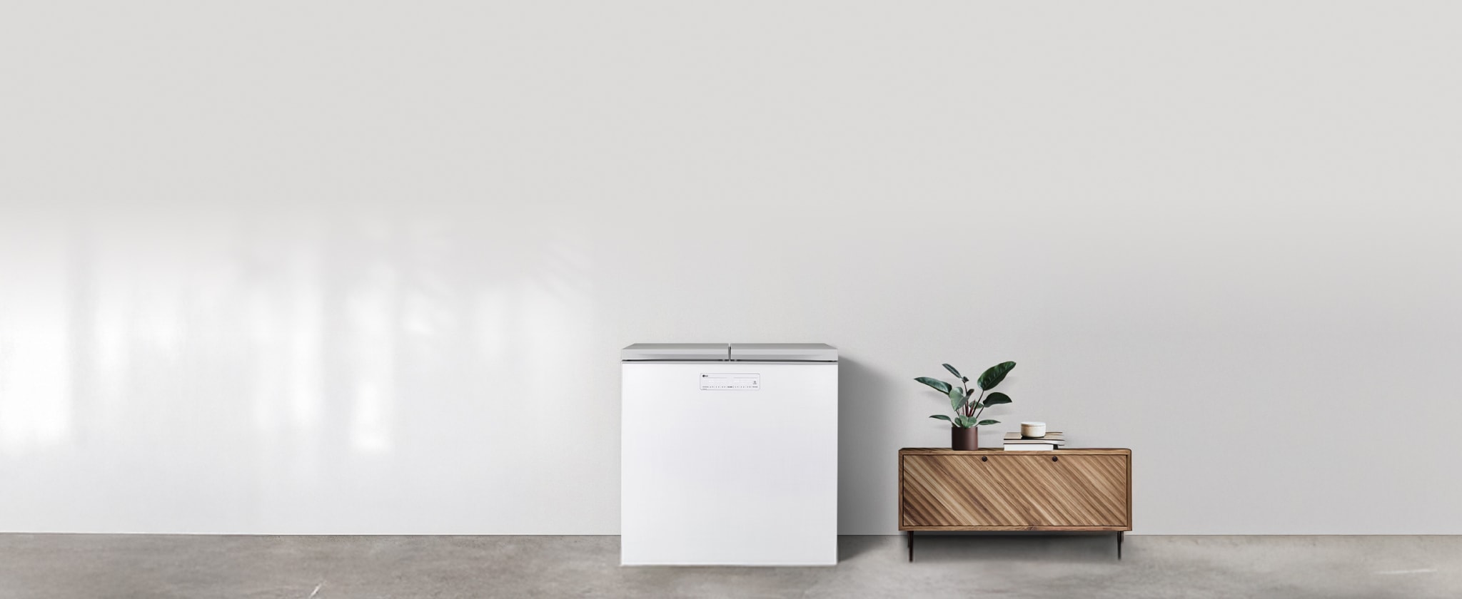 밝고 심플한 공간에 연출된 미니멀 인테리어로, 왼쪽에는 상부 개폐형 화이트 김치냉장고가 배치되어 있습니다. 오른쪽에는 사선 무늬가 돋보이는 원목 수납장이 놓여 있으며, 그 위에는 초록 식물 화분과 컵, 책이 정돈되어 있습니다.