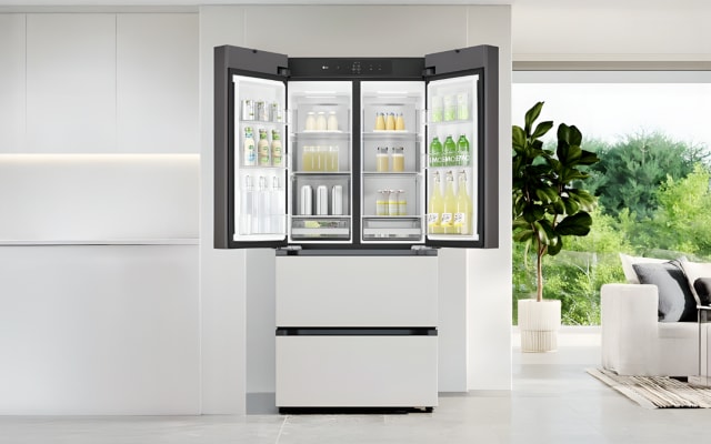 중앙에는 도어가 활짝 열린 매트 화이트 프렌치도어 김치냉장고가 설치되어 있습니다. 냉장고 내부에는 각종 음료와 유제품이 정돈되어 있으며, 투명한 선반 구조로 내용물이 한눈에 잘 보이도록 구성되어 있습니다. 오른쪽에는 초록 식물과 함께 자연 풍경이 보이는 대형 창이 있어 공간 전체에 밝고 탁 트인 분위기를 더합니다.