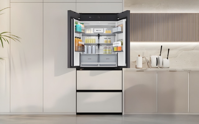 베이지 톤의 주방 공간으로, 중앙에는 도어가 열린 매트 화이트 프렌치도어 김치냉장고가 설치되어 있습니다. 냉장고 내부에는 음료와 유제품이 카테고리별로 정돈되어 있으며, 투명한 선반과 서랍 구조로 내용물이 깔끔하게 진열되어 있습니다. 왼쪽에는 대형 화분이 놓여 자연의 요소를 더하고, 오른쪽에는 간결한 조리대와 수납장이 일체감 있게 구성되어 모던하고 미니멀한 분위기를 연출합니다.