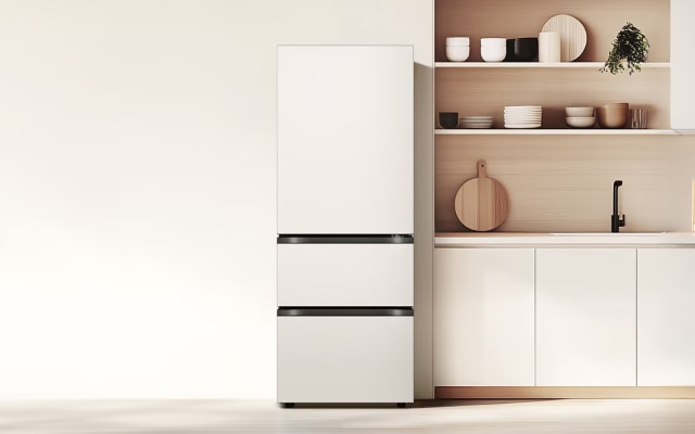 우드 톤과 화이트가 조화를 이루는 미니멀한 주방 공간으로, 왼쪽에는 매트 화이트 하단 서랍형 김치냉장고가 설치되어 있습니다. 오른쪽에는 오픈 선반과 수납장이 이어지며, 선반 위에는 식기와 소품이 정갈하게 정리되어 있습니다. 조리대에는 나무 도마, 허브 화분, 과일 등이 배치되어 있고, 밝은 톤의 벽과 바닥이 어우러져 전체적으로 따뜻하고 차분한 분위기를 자아냅니다.