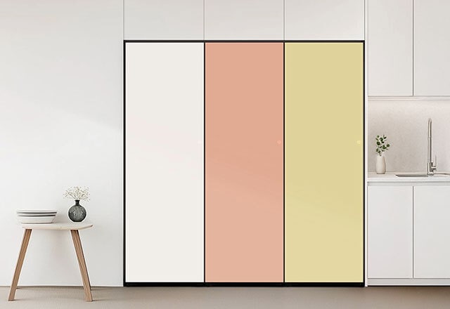 부드러운 베이지 톤과 화이트가 조화를 이루는 미니멀한 주방 공간으로, 중앙에는 매트 화이트와 메탈릭 베이지 컬러가 어우러진 빌트인 김치냉장고가 설치되어 있습니다. 왼쪽에는 대형 창과 초록 식물 화분이 자연 채광과 생기를 더하며, 오른쪽에는 베이지 수납장과 스테인리스 싱크대, 간결한 그릇들이 정돈되어 있어 전체적으로 세련되고 조화로운 분위기를 연출합니다.