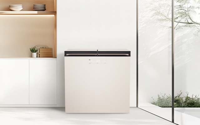 밝고 깔끔한 미니멀 인테리어 공간으로, 중앙에는 상부 개폐형 매트 크림 화이트 김치냉장고가 배치되어 있습니다. 왼쪽에는 화이트 하부장 위에 식물 화분과 나무 도마가 정돈되어 있으며, 오른쪽의 전면 유리 통창을 통해 햇빛과 정원이 어우러져 자연스러운 분위기를 연출합니다.