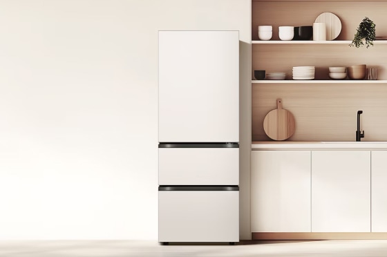 미니멀한 디자인의 주방 공간으로, 왼쪽에는 매트 화이트 컬러의 하단 서랍형 김치냉장고가 배치되어 있습니다. 오른쪽에는 연한 우드 톤의 오픈 선반과 하부장이 구성되어 있으며, 선반 위에는 식기류와 소품이 정돈되어 있고 나무 도마와 화분이 따뜻한 분위기를 더합니다.