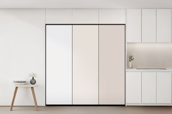 깔끔한 올화이트 인테리어 공간 중앙에는 빌트인 3도어 김치냉장고가 설치되어 있으며, 좌측부터 화이트, 크림, 베이지 톤의 패널이 세련된 색상 조화를 이룹니다. 오른쪽에는 간결한 싱크대와 화병이 놓인 조리 공간이, 왼쪽에는 작은 원형 테이블과 화병이 포인트로 배치되어 전체적으로 모던하고 정돈된 분위기를 자아냅니다.