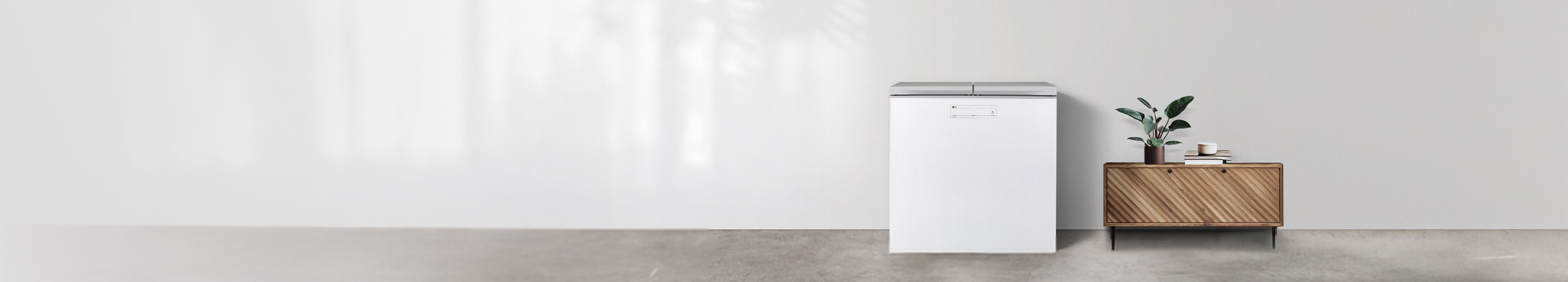 밝고 심플한 공간에 연출된 미니멀 인테리어로, 왼쪽에는 상부 개폐형 화이트 김치냉장고가 배치되어 있습니다. 오른쪽에는 사선 무늬가 돋보이는 원목 수납장이 놓여 있으며, 그 위에는 초록 식물 화분과 컵, 책이 정돈되어 있습니다.