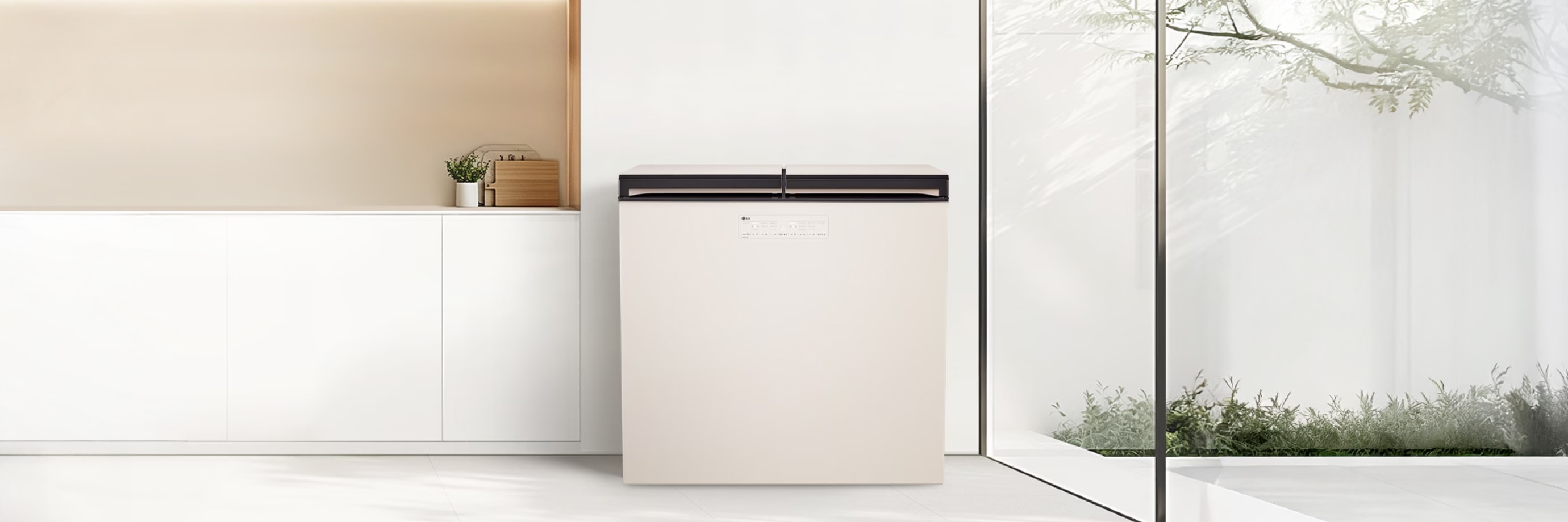 밝고 깔끔한 미니멀 인테리어 공간으로, 중앙에는 상부 개폐형 매트 크림 화이트 김치냉장고가 배치되어 있습니다. 왼쪽에는 화이트 하부장 위에 식물 화분과 나무 도마가 정돈되어 있으며, 오른쪽의 전면 유리 통창을 통해 햇빛과 정원이 어우러져 자연스러운 분위기를 연출합니다.