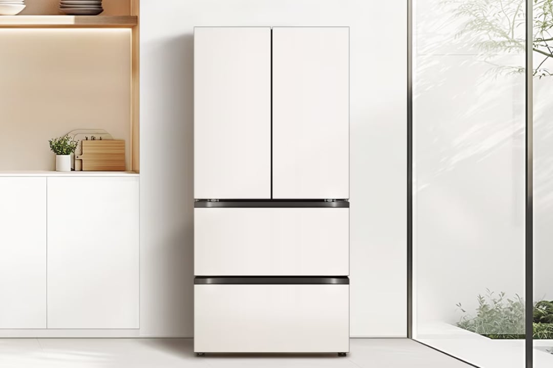 깔끔하고 밝은 주방 공간으로, 중앙에는 매트 화이트 컬러의 하단 서랍형 프렌치도어 김치냉장고가 설치되어 있습니다. 왼쪽에는 화이트 하부장과 오픈 선반이 구성되어 있으며, 식물 화분과 나무 도마가 정돈되어 따뜻한 분위기를 더합니다. 오른쪽 전면 유리창을 통해 자연 채광이 들어와 공간에 개방감을 더합니다.