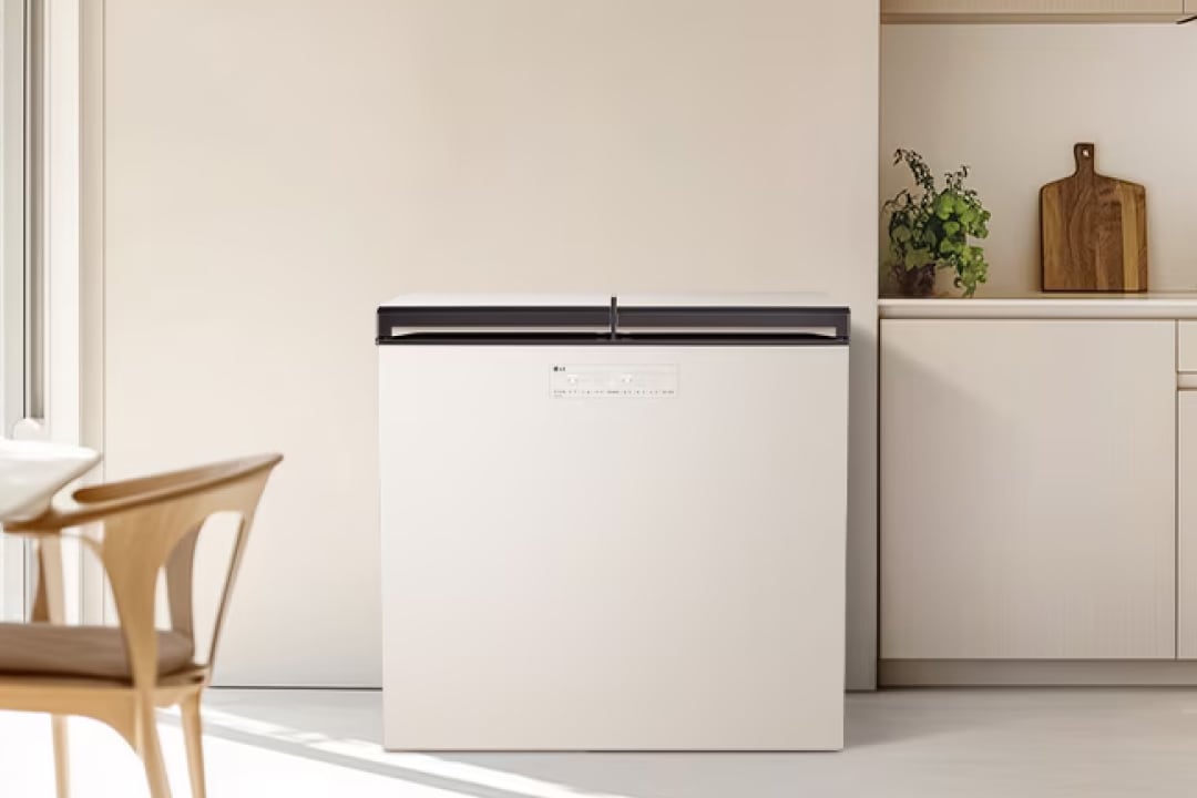 따뜻한 자연광이 스며드는 미니멀한 주방 공간으로, 중앙에는 상부 개폐형 매트 화이트 김치냉장고가 배치되어 있습니다. 오른쪽에는 화이트 하부장과 오픈 선반이 구성되어 있으며, 선반 위의 초록 식물과 나무 도마가 공간에 생기와 온기를 더합니다. 왼쪽에는 원목 의자가 놓인 식탁 공간이 일부 보이며, 전체적으로 차분하고 따뜻한 분위기를 자아냅니다.