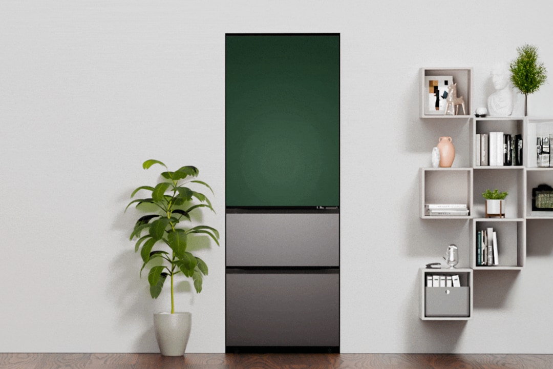 중앙에는 딥 그린 도어와 다크 그레이 서랍이 조합된 하단 서랍형 김치냉장고가 설치되어 있습니다. 왼쪽에는 싱그러운 녹색 식물 화분이 놓여 있고, 오른쪽 벽면에는 소품과 책이 정돈된 오픈형 큐브 선반이 구성되어 있습니다.