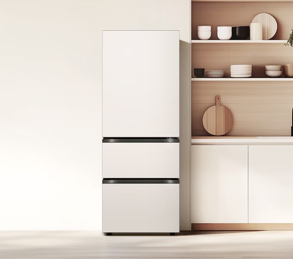 미니멀한 디자인의 주방 공간으로, 왼쪽에는 매트 화이트 컬러의 하단 서랍형 김치냉장고가 배치되어 있습니다. 오른쪽에는 연한 우드 톤의 오픈 선반과 하부장이 구성되어 있으며, 선반 위에는 식기류와 소품이 정돈되어 있고 나무 도마와 화분이 따뜻한 분위기를 더합니다.