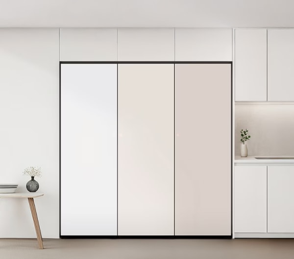 깔끔한 올화이트 인테리어 공간 중앙에는 빌트인 3도어 김치냉장고가 설치되어 있으며, 좌측부터 화이트, 크림, 베이지 톤의 패널이 세련된 색상 조화를 이룹니다. 오른쪽에는 간결한 싱크대와 화병이 놓인 조리 공간이, 왼쪽에는 작은 원형 테이블과 화병이 포인트로 배치되어 전체적으로 모던하고 정돈된 분위기를 자아냅니다.