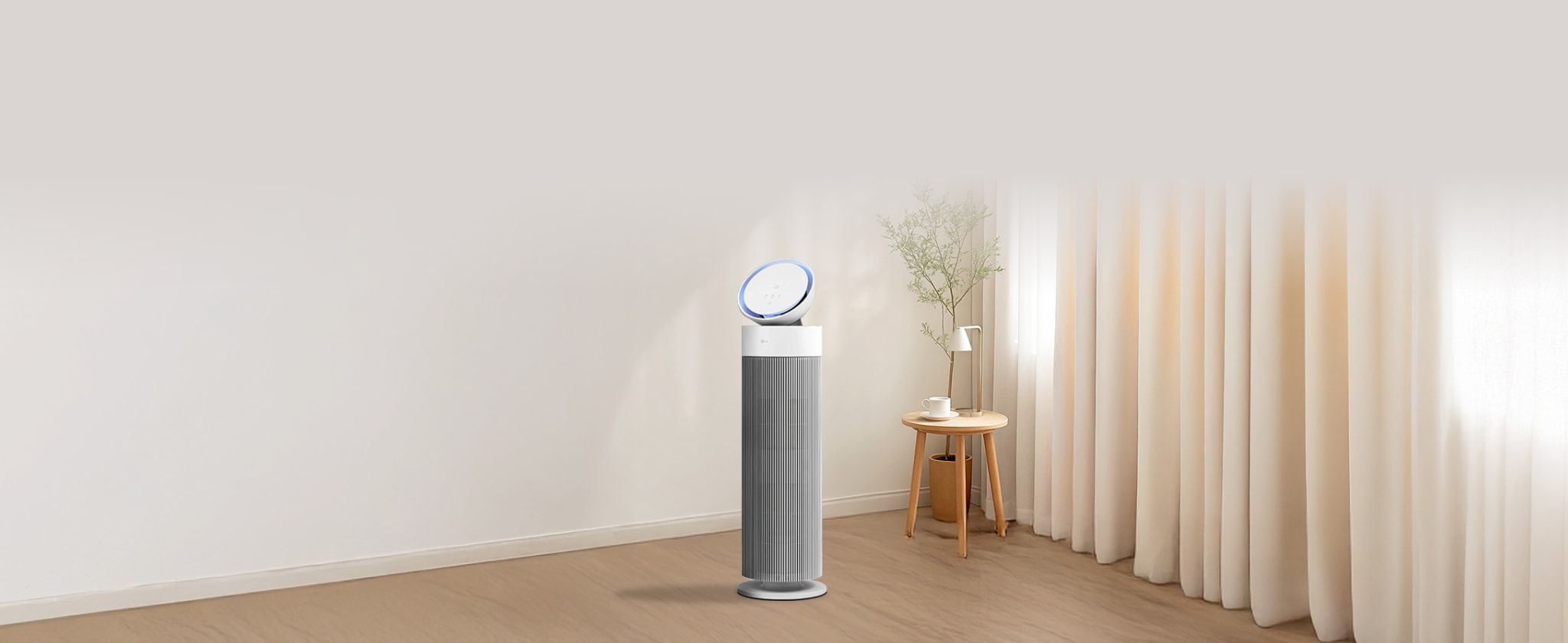 밝고 현대적인 거실에서 LG 퓨리케어 오브제컬렉션 에어로부스터가 중앙에 위치하며, 세련된 원통형 디자인과 상단의 기울어진 원반형 디스플레이가 특징입니다. 옆에는 화분으로 장식되어 있고, 뒤로는 창문이 햇빛을 받아 밝은 분위기를 조성합니다. 전체적으로 미니멀하고 깨끗한 공간을 연출합니다.