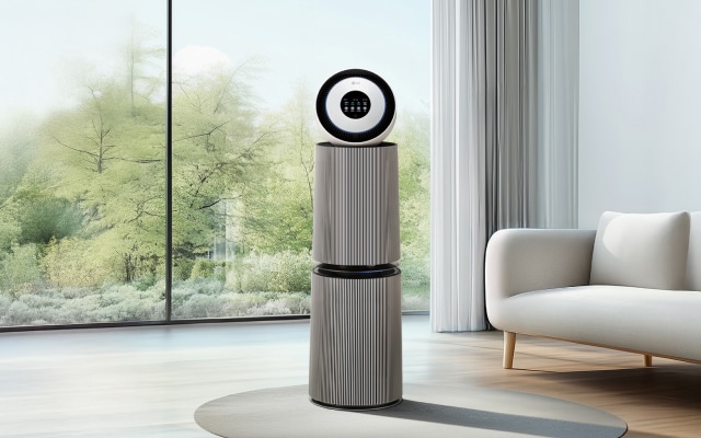 차분하고 깔끔한 거실 공간에 공기청정기가 놓여 있습니다. 원형 헤드와 세로 결 무늬가 돋보이는 본체 디자인이 세련된 분위기를 더하며, 옆에는 화이트 소파와 커튼이 어우러져 편안한 공간감을 연출합니다. 전면 유리창 너머로는 푸른 숲이 펼쳐져 자연과 조화로운 인테리어를 완성합니다.