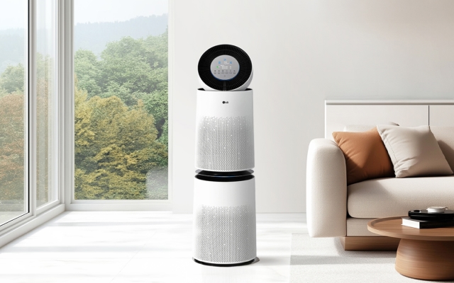 따뜻한 톤의 거실 공간에 공기청정기가 배치되어 있습니다. 화이트 컬러와 미세한 패턴이 조화를 이루는 본체 디자인이 모던한 분위기를 연출하며, 옆에는 베이지 톤의 소파와 라운드 테이블, 러그가 어우러져 아늑하고 편안한 공간감을 자아냅니다.