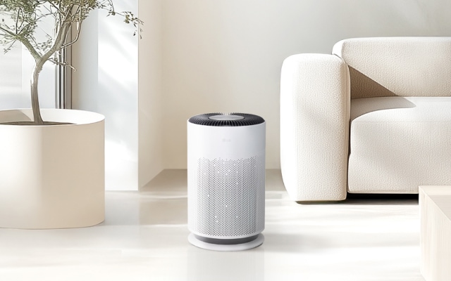 깔끔하고 따뜻한 분위기의 실내 공간에 화이트 컬러의 원형 공기청정기가 놓여 있습니다. 미니멀한 디자인이 인테리어에 자연스럽게 어우러지며, 옆의 메탈 프레임 수납장과 조화를 이루어 모던한 감성을 더합니다. 창문을 통해 들어오는 자연광이 공간을 부드럽게 밝혀 쾌적하고 정돈된 분위기를 연출합니다.