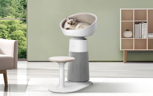 넓은 창으로 햇살이 들어오는 밝은 거실에 캣타워형 공기청정기가 놓여 있으며, 상단 쿠션 베드에는 고양이가 편안하게 누워 있습니다. 곁에는 고양이 점프용 스텝이 함께 배치되어 있어 반려동물의 휴식과 이동을 돕습니다. 전체적으로 따뜻하고 아늑한 분위기가 느껴지는 공간입니다.
