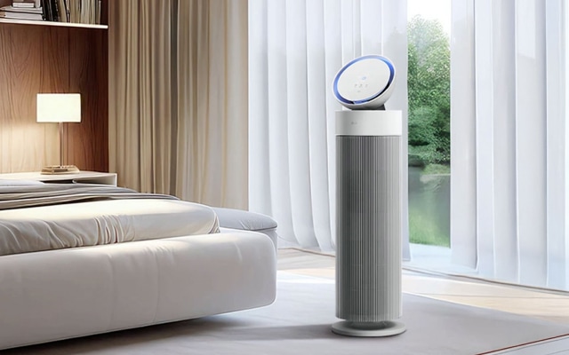 침실 환경에서 원통형 디자인의 LG 퓨리케어 오브제컬렉션 에어로부스터가 보입니다. 상단에는 원반형 디스플레이가 있고, 하단은 회색 세로 줄무늬가 있는 디자인입니다. 옆에는 흰색 침대가 놓여 있으며 뒤쪽 배경에는 질감 있는 흰색 벽면이 보입니다.