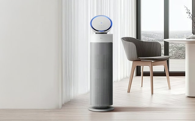 채광이 좋은 실내에 LG 퓨리케어 오브제컬렉션 에어로부스터가 놓여있습니다. 옆에는 식탁, 의자가 배치되어 있습니다. 공기 청정기는 원통형 몸체와 파란색 링 조명이 있는 원형 디스플레이로 구성되어 있으며, 식탁은 원형 상판과 기둥 모양 다리를 가지고 있고, 의자는 패브릭 소재의 등받이와 나무 다리를 가지고 있습니다.