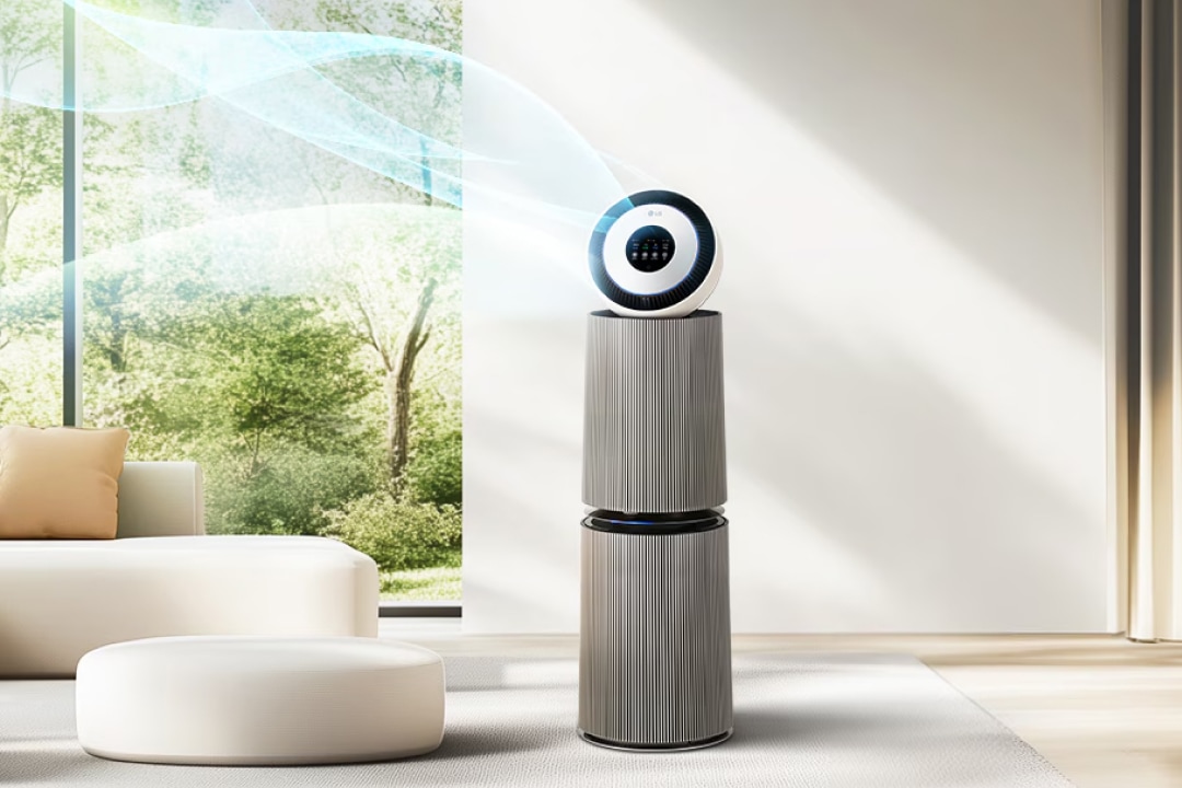 세련된 실내 공간에 공기청정기가 놓여 있습니다. 공기청정기에서 맑은 공기가 나오는 모습이 보입니다. 창문 너머로 푸른 나무들이 보이며, 실내에는 베이지색 소파와 둥근 쿠션이 있습니다.