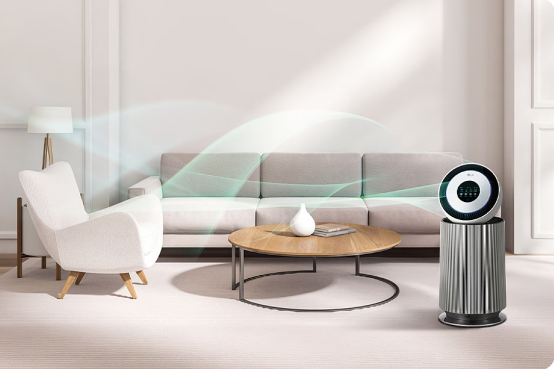 세련된 실내 공간에 공기청정기가 놓여 있습니다. 공기청정기는 원통형 모듈로 구성되어 있으며, 상단에 원형 디스플레이가 있습니다. 디스플레이에는 LG라는 로고가 보입니다. 주변에는 큰 창문을 통해 녹색 나무와 도시 풍경이 보이며, 흰색 소파와 낮은 테이블이 배치되어 있습니다. 테이블 위에는 책과 소품들이 놓여 있습니다.