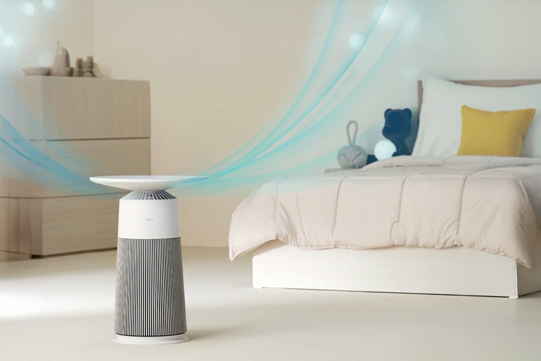 침실 환경에 공기청정기가 놓여있습니다. 공기청정기에서 맑은 공기가 나오는 모습이 보입니다. 배경에는 단순하고 현대적인 나무 서랍장과 침대가 있고, 침대에는 밝은 색상의 베개와 쿠션이 놓여 평온하고 따뜻한 실내 분위기를 더합니다.