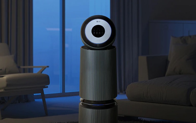 어두운 거실에서 작동 중인 LG 공기청정기. 원형 디스플레이에 불이 켜져 있으며, 주변에는 창문과 소파, 의자가 보임. 조명이 없는 밤 시간의 분위기를 연출.