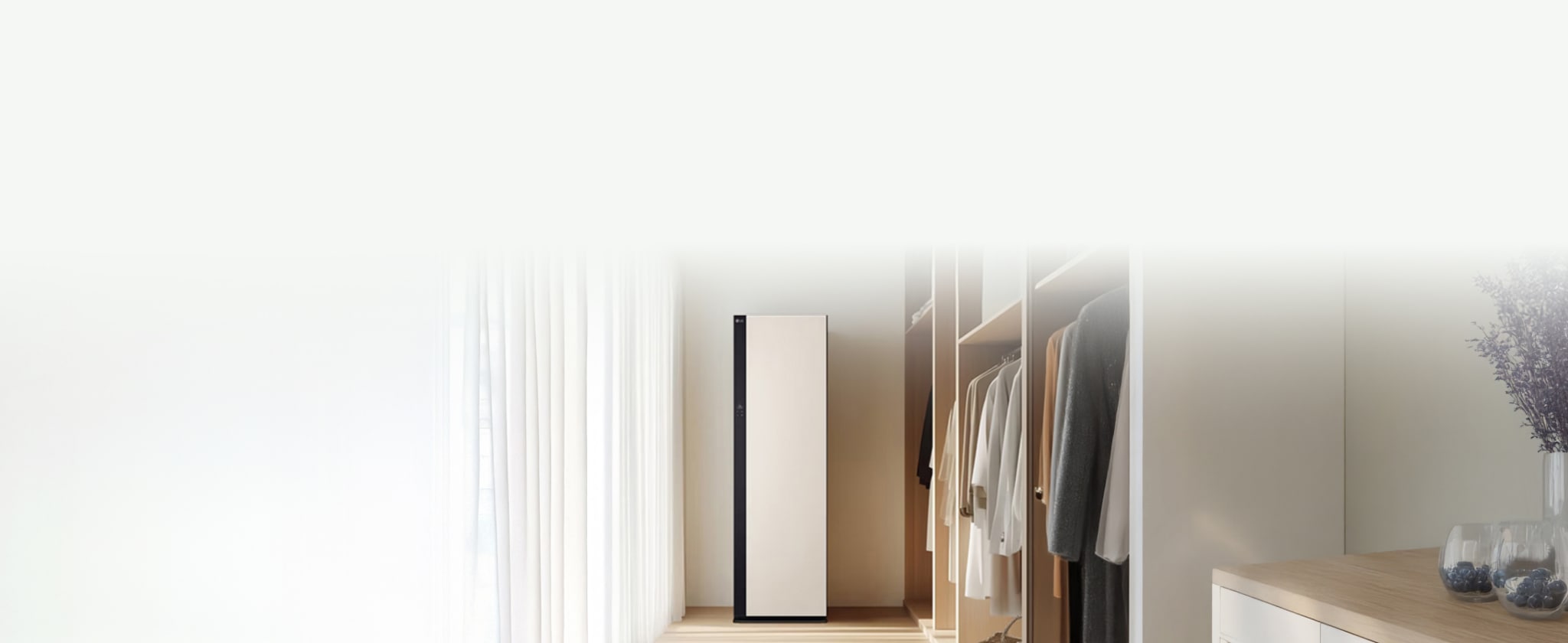 따뜻한 드레스룸 공간에 옷장 속 정갈하게 걸려있는 옷가지들이 있습니다. 가운데에는 밝은 색상의 스타일러가 놓여져있고, 왼쪽에 아이보리색의 커튼이 함께 배치되어있습니다.