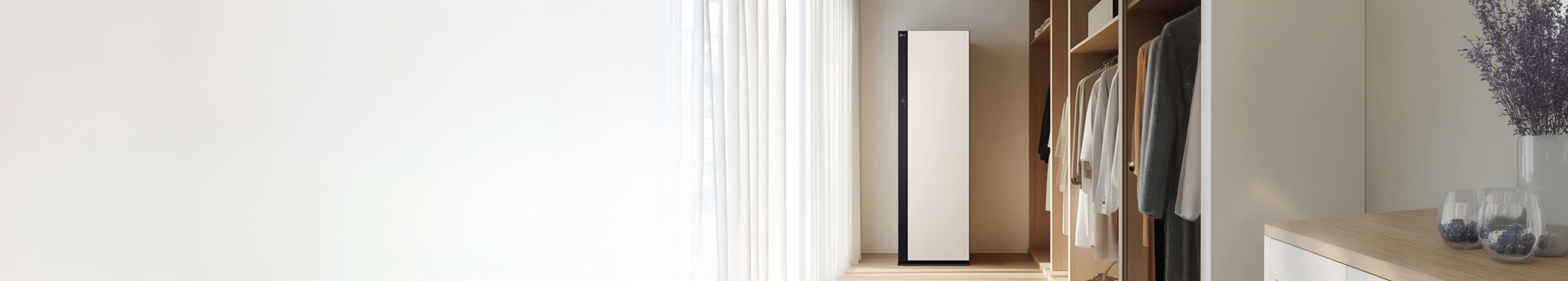 따뜻한 드레스룸 공간에 옷장 속 정갈하게 걸려있는 옷가지들이 있습니다. 가운데에는 밝은 색상의 스타일러가 놓여져있고, 왼쪽에 아이보리색의 커튼이 함께 배치되어있습니다.