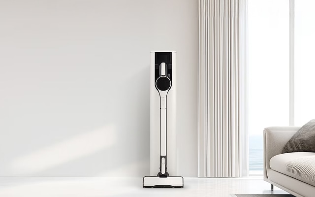 밝은 거실 배경에 흰색 무선 청소기가 세워져 있고, 옆에는 베이지색 수납장과 회색 꽃병, 흰색 소파가 배치되어 있습니다.