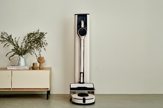 아이보리색 벽을 배경으로, 흰색과 검은색의 LG 무선 청소기와 로봇 청소기가 충전 거치대와 함께 놓여 있습니다. 충전 거치대 옆에는 나무 장식품이 올려진 크림색과 나무색의 낮은 서랍장이 부분적으로 보입니다.