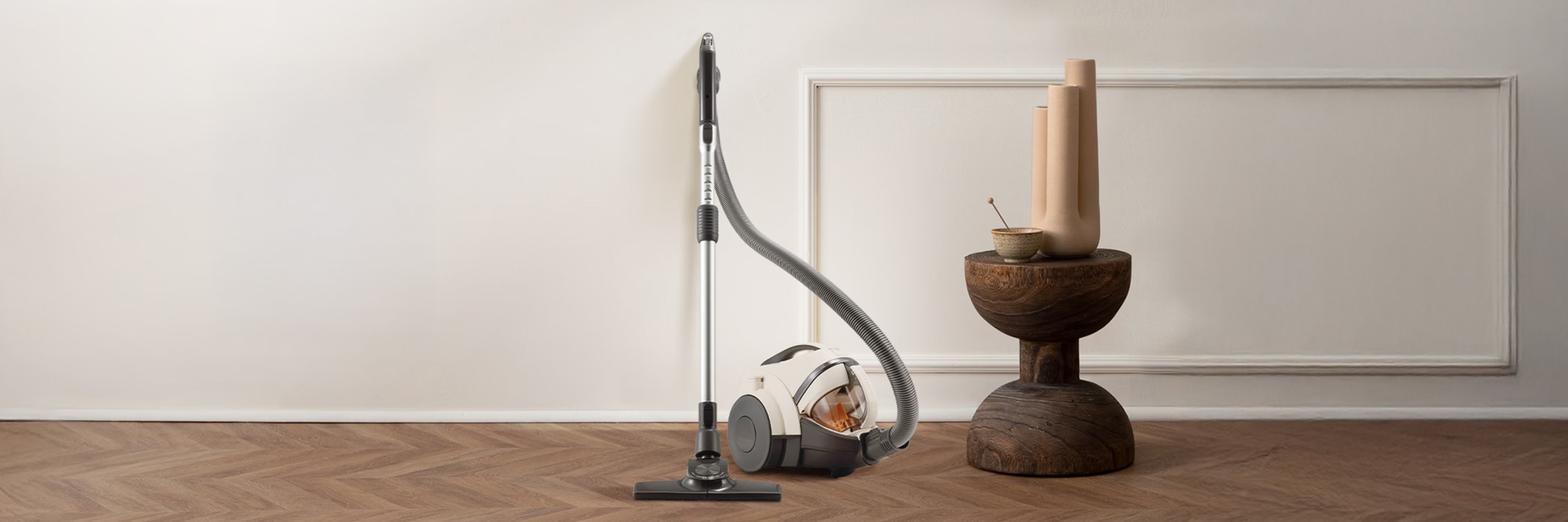 헤링본 패턴의 나무 바닥 위에 흰색과 회색의 유선 진공청소기가 놓여 있습니다. 옆에는 나무 받침대 위에 도자기 꽃병과 작은 그릇 장식이 함께 놓여 있고 배경은 흰색 벽과 몰딩으로 장식되어 있습니다.