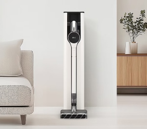 미니멀한 거실 공간에 화이트 컬러의 무선청소기가 스탠드형 충전 거치대에 깔끔하게 보관되어 있어 정돈된 인테리어와 조화를 이루고 있습니다.