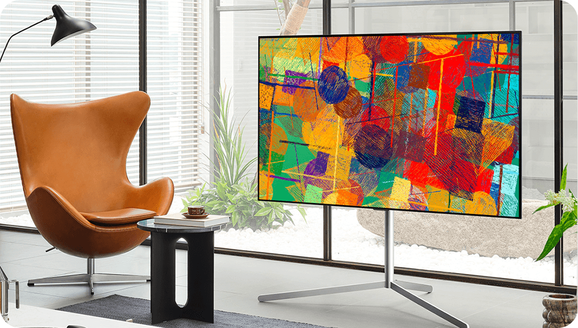 밝은 거실에 설치된 스탠드형 LG TV 화면에 다채로운 색상의 추상화가 표시되어 있으며, 옆에는 갈색 가죽 의자와 작은 원형 테이블이 배치된 모습.