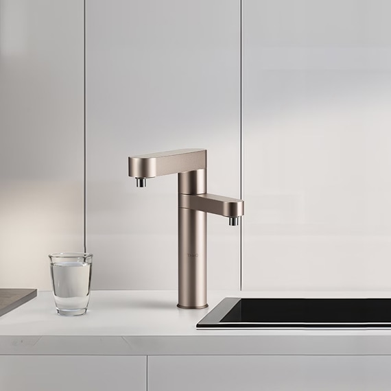 밝은 조명이 비추는 따뜻한 분위기의 인테리어 공간 한가운데, 브론즈 톤의 LG 정수기가 놓여 있으며, 그 아래에는 투명 유리컵에 물이 채워져 있음.