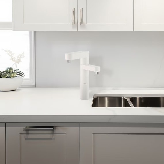 화이트 색상의 듀얼 정수기가 빌트인 설치되어 있는 주방 공간. 싱크대 옆에 설치되어 있으며, 주방 상부장은 화이트, 하부장은 그레이 색상. 왼쪽에는 창문 앞에 화분이 하나 놓여져 있다.