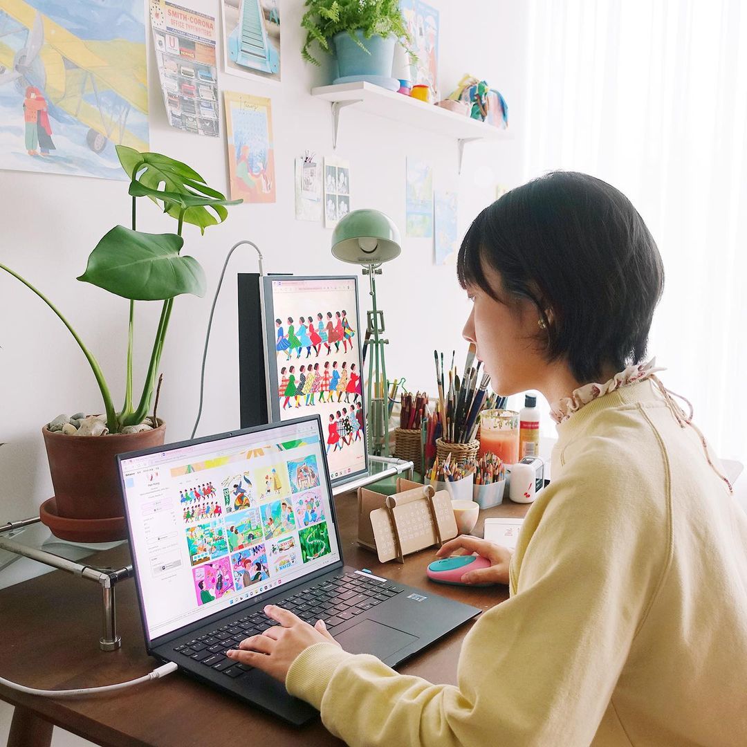 LG 그램 노트북과 세로로 세운 그램+view 포터블 모니터를 사용해 그래픽 작업 중인 여성. 책상 위에는 식물, 그림 도구, 조명이 놓여 있으며, 벽에는 다양한 일러스트와 장식이 걸려 있음.