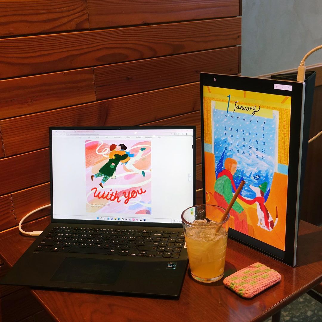 나무 테이블 위 LG 그램 노트북과 세로로 세운 그램+view 포터블 모니터가 놓여 있으며, 각각 일러스트 작업물과 캘린더가 표시됨. 옆에는 아이스 음료와 뜨개질 된 코스터가 놓여 있음.