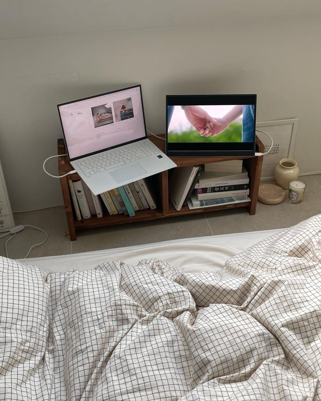 침대 앞 나무 책장 위 LG 그램 노트북과 그램+view 포터블 모니터가 놓여 있음. 노트북에는 웹사이트, 모니터에는 손을 맞잡은 영상이 표시됨. 책과 촛불, 도자기 장식이 어우러져 아늑한 분위기를 연출함.