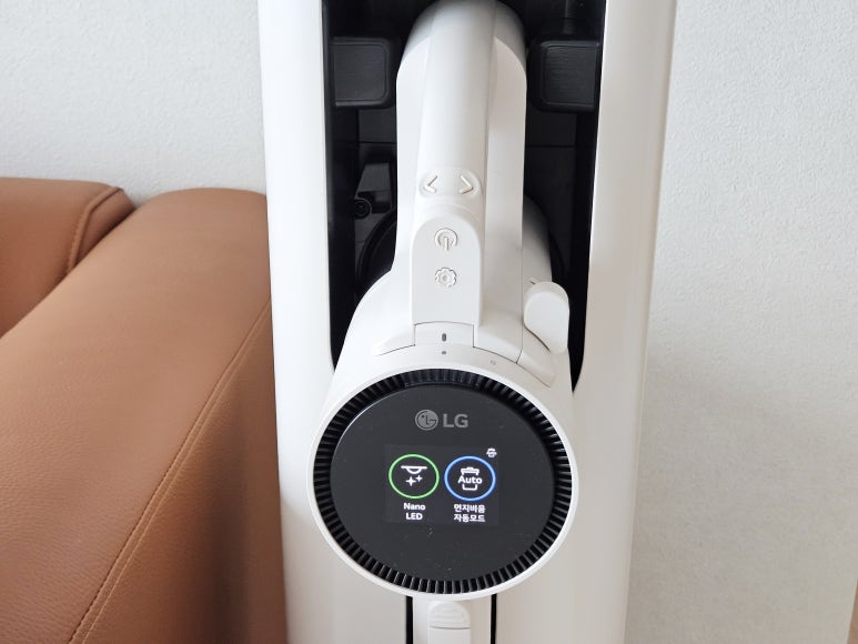 코드제로_a9s가_충전_거치대에_세워져_있으며,_디스플레이_화면에_nano_led와_먼지비움_자동모드가_표시됨._왼쪽에는_갈색_가죽_소파가_있음.
