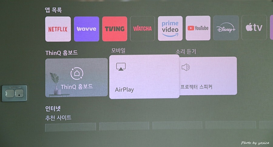 프로젝터의 홈 화면임. 넷플릭스, 웨이브, 티빙 등 주요 스트리밍 앱과 AirPlay, 소리 설정 등 다양한 기능이 표시되어 있음. 직관적이고 깔끔한 UI가 사용자 편의를 높임.