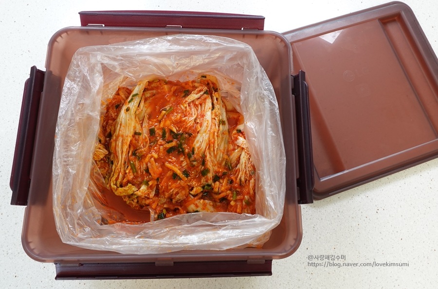조리대_위에_올려진_갈색_김치통,_뚜껑이_열려_있음._투명_비닐_안에_양념된_배추김치가_담겨_있음.김조리대_위에_올려진_갈색_김치통,_뚜껑이_열려_있음._투명_비닐_안에_양념된_배추김치가_담겨_있음.