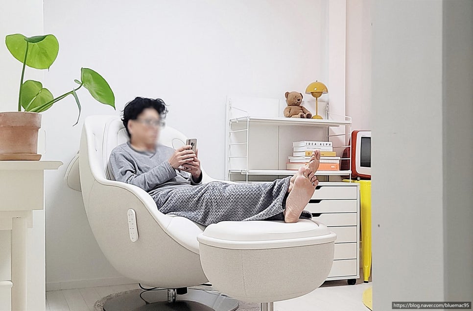 흰색_안마의자에_앉아_발발침에_발을_올리고_스마트폰을_사용하는_남성._옆에는_푸른_잎_화분이_배치돼_있고,_뒤로는_책과_장식품들이_정리된_선반이_보임.
